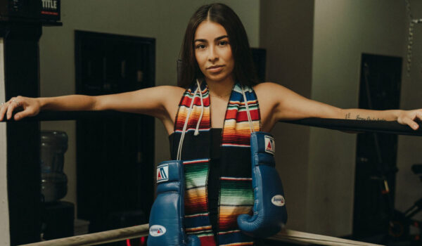 A woman leans against the ropes of a boxing ring. She has boxing gloves hanging from her neck as well as colorful graduation stoles.