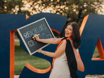 A woman in a tan dress holds a chalk board with the boxes checked off for "bachelor's" and "master's"