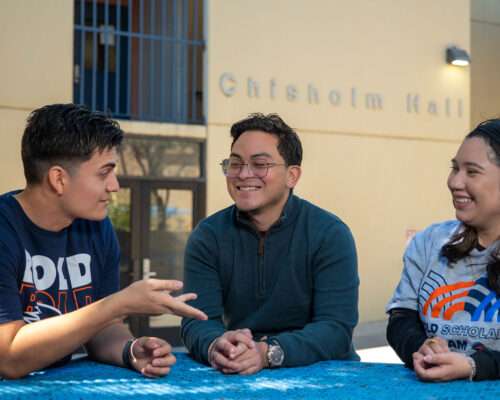 Three people sit at a table table outdoors smiling and talking together.