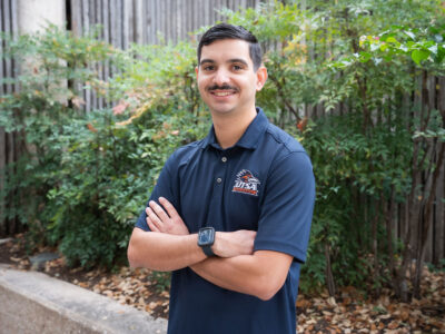 A man in a blue polo folds his arms and smiles.