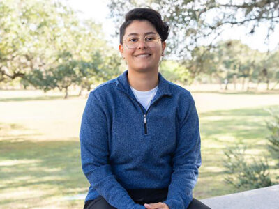 A woman in a bluw collard sweater and glasses sits for a photo.