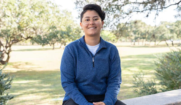 A woman in a bluw collard sweater and glasses sits for a photo.