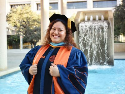 A woman with brown curly hair stands in front of a water fountain dressed in commencement regalia.
