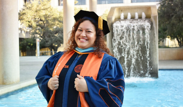 A woman with brown curly hair stands in front of a water fountain dressed in commencement regalia.