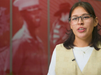 A woman with glasses, a white shirt and yellow checked vest, stands outside of the Frost Tower, which houses the Institute of Texan Cultures.