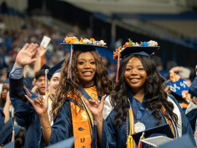 Two woman stand in a middle of a crowed as they are dressed in a blue graduation cap and gown.