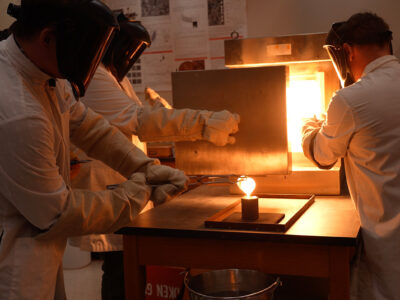 People in white lab coats and protective gear hold rocks over a flame to melt them.