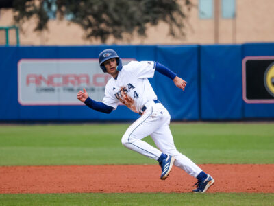 A young man in a baseball uniform runs to the left on a baseball field.