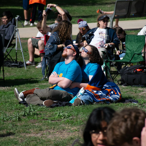 A couple sits on a lawn hugging each other as they wear solar eclipse glasses.