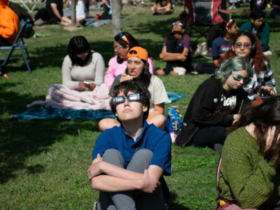 A community member sits on a lawn with eclipse glasses on.