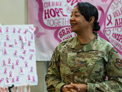 A woman in a military uniform looks off to the side. There are Breast Cancer Awareness Month posters behind her.