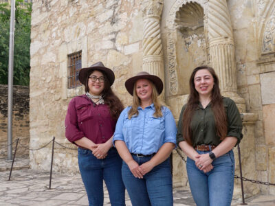 Three women, two of them in hats, stand side by side in front of a building called The Alamo.