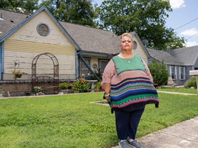 A lady stands on her front lawn and looks off to the side.