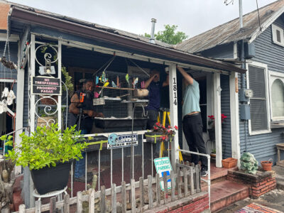 Three individuals stand on the front porch of a house as two of them are hanging something from the cieling.